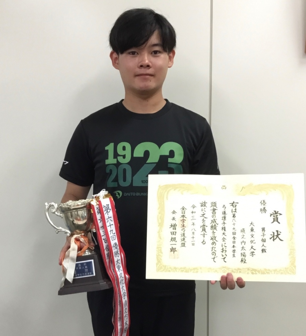 弓道部の須之内太陽選手(法律学科1年)が第69回全日本学生弓道選手権大会男子個人戦で優勝し、100周年記念Ｔシャツを着用してスポーツ振興センターに優勝報告に来てくれました。
遠征先の宿舎でも弓道部員は100周年記念Ｔシャツを着用していたとのことです。